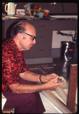 A photographic slide of Don E. Crabtree flintknapping with obsidian while seated inside.