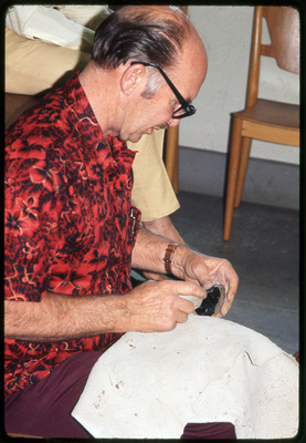A photographic slide of Don E. Crabtree flintknapping with obsidian while seated inside.