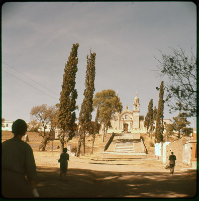 A photographic slide displaying the front of an ornate structure, possibily a church. A stairway leading to the front door is lined with trees, and a few people stand in the foreground facing away. The entire slide is tinted red.