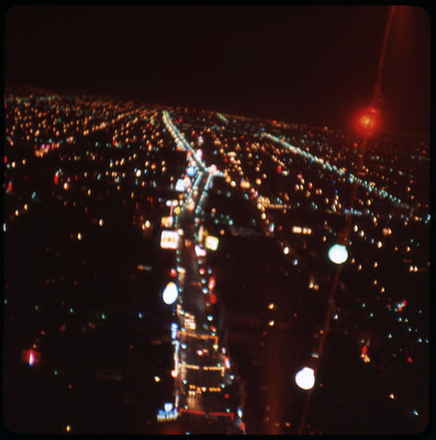 A photographic slide displaying an aerial view of a cityscape at night. Bright, blurry, multicolored lights dot the scene.