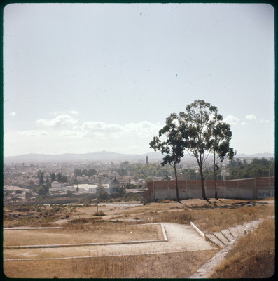 A photographic slide displaying an overview of the outskirts of a city. There is a few small trees in the foreground, a road behind them, and a townscape behind that. The entire slide is tinted red.