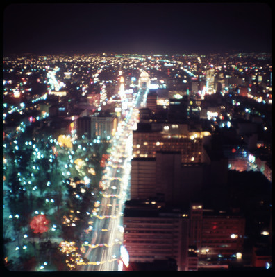 A photographic slide displaying an aerial view of a cityscape at night. Bright, blurry, multicolored lights dot the scene.
