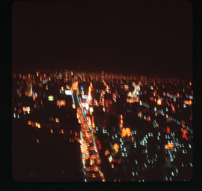 A photographic slide displaying an aerial view of a cityscape at night. Bright, blurry, multicolored lights dot the scene.