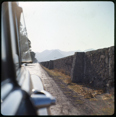 A photographic slide displaying a long stone wall as seen from the side of a vehicle. Behind the wall is a large mountain range.