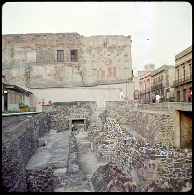 A photographic slide displaying an old brick building. Painted words on the side of the building have since faded. Below it is a set of stone, walled pathways.