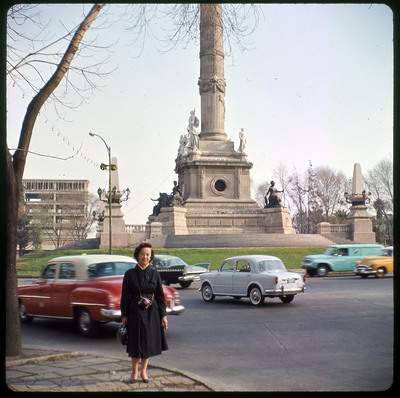 A photographic slide displaying Evelyn Crabtree standing in front of a tall stone monument. The monument is a large, tall column surrounded by various statues. The top of the column is cut off by the frame.