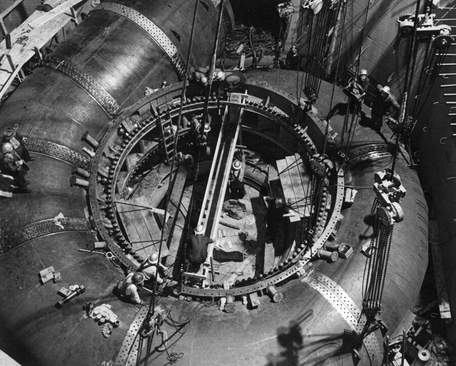 The 190-ton scroll case for 103,000 horsepower Allis-Chalmers hydro turbine unit being installed in the west powerhouse at Grand Coulee Dam. This unit consists of several radial and on straight section of welded plate steel riveted with double butt straps. One half of combined unit is lifted by an overhead crane and moved into position. Before being poured in concrete, the scroll case is given hydraulic test of 310 pounds to the square inch.  This scroll case, designed for use at Shasta Dam in northern California, is temporarily installed at Grand Coulee Dam by order of the War Production Board, as construction at Shasta Dam had not progressed sufficiently to make immediate use of turbine at that point. With the waters of the mighty Columbia River as a driving force, this turbine now installed is spinning a giant generator at a rate of 138.5 revolutions per minute, producing more than 75,000 kilowatts of electrical energy.