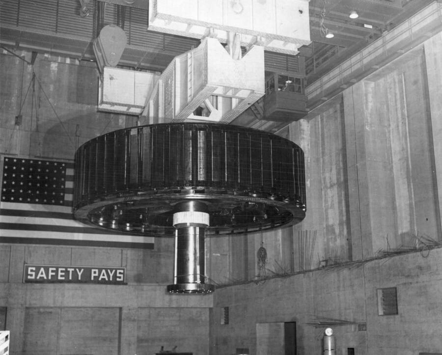Col. Basin Proj., Grand Coulee Dam.--Rotor for main generator unit L-6 in the west powerhouse at Grand Coulee Dam being carried to final position by two overhead travelling cranes using an equalizing beam. The rotor, 31 feet in diameter, weighing 535 tons, rotates at a speed of 120 revolutions per minute within stator, and develops 106,000 kilowatts of electrical energy.