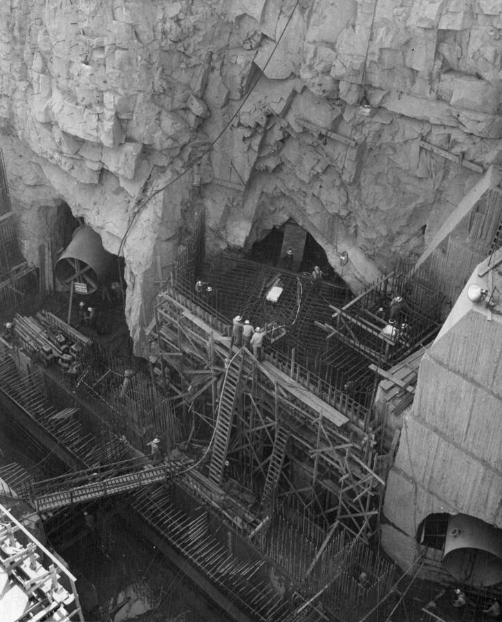 Construction of the Pumping Plant Structure, Grand Coulee Dam, Washington. Installation of pumping unit P-2 discharge pipe, showing the concrete form around the outlet tunnel, placement of reinforcing steel and in the center close to the concrete form is a square opening for the block out which is on top of the discharge pipe under the steel matt. This block out extends upward on an angle from the end of the steep matt to the top of the tunnel, part of it can be seen in the center background, it continues on into the tunnel and will be used to run pumpcrete pipes for the pouring of concrete around the discharge pipe. The vertical steel bars on the steel matt (right) are for the elevator block out. Discharge pipes for pumping units P-1 and P-3 have been placed in position and are shown at the right and left of P-2 installation.
