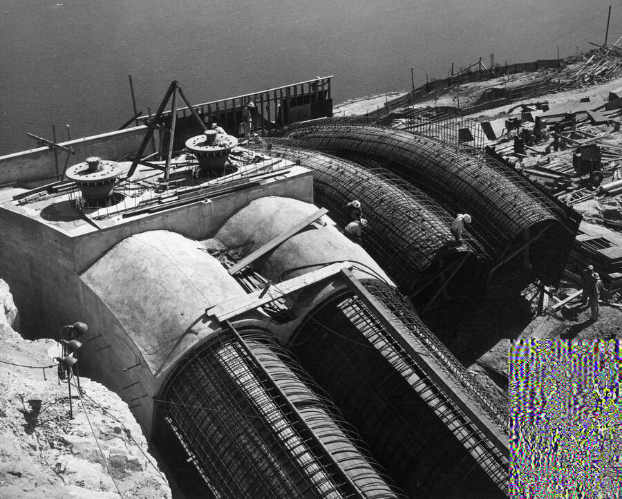 Specifications 2329. Morrison-Knudsen Company, Inc., and Peter Kiewit Sons' Company. Progress view of the pump-discharge outlet structure showing the status of construction of the crest bends of P-1 to P-4 discharge pipes. Pipes P-1 and P-2 have been encased with concrete, with the steel pipe ending at this point as can be seen in lines P-3 to P-4 around which reinforcing steel is being placed. The pipes are to extend from this point with round reinforced concrete tubes as can be seen in the foreground where P-1 and P-2 are formed and the placing of reinforcing steel started. Note the two siphon breakers installed on P-1 and P-2 crest bends.