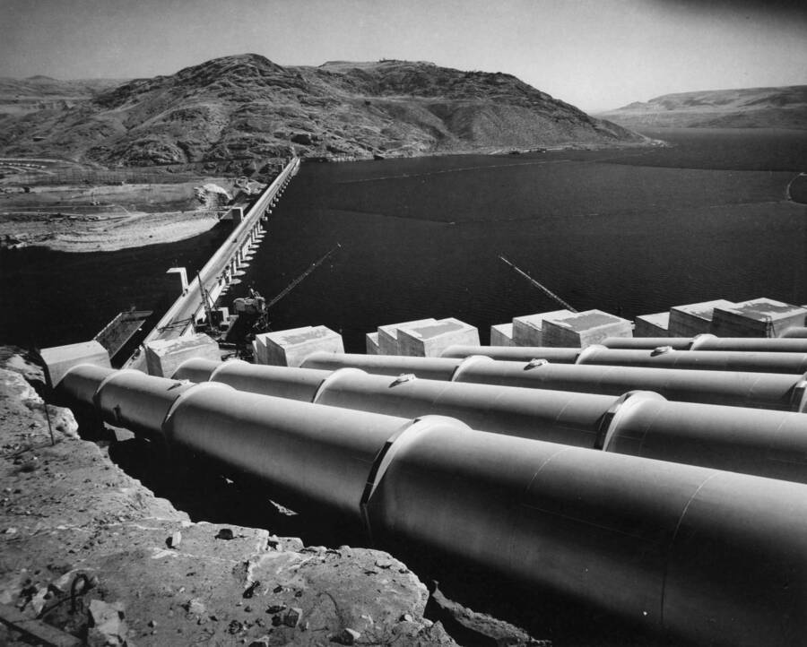 The exposed portions of the pumping plant discharge pipes gleam brightly in the sun with their coat of aluminum paint. Symbolic of the entire Columbia Basin Project--this picture shows Coulee Dam holding back the waters of the Columbia River forming Lake Roosevelt. The water there held, is now awaiting only the pump installations to start the water flowing through these pipes and to the thirsty acres of the project 60 to 150 miles south of the dam.
