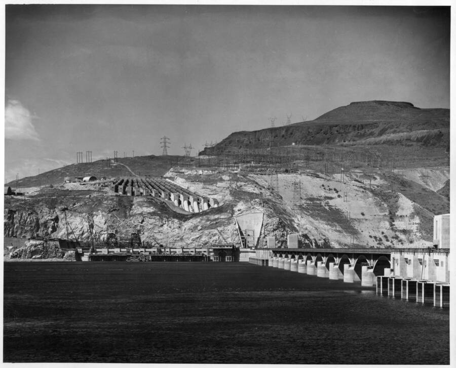 View of the west abutment from the east parking lot showing the discharge pipes, dam pumping plant and Coulee Dam.