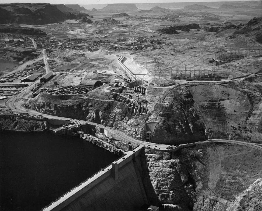 Specifications 2329. Contractor: Morrison-Knudsen Company, Inc. and Peter Kiewit Sons' Company. This aerial view of the Grand Coulee Dam area shows the status of progress on the construction of Grand Coulee pumping plant and feeder canal. The pumping plant is being constructed behind the 600-foot long wing dam extending to the left from near the west abutment end of Grand Coulee Dam. The world's largest pumps will be installed in the pumping plant at a level of 120 feet below the roadway elevation. Each pump will lift water 280 feet from Roosevelt Lake and will discharge it through a steel pipe 12 feet in diameter and about 680 feet in length. Twelve tunnels, from 487 to 530 feet in length, will carry the pipes under the highway and up through the granite canyon wall. Upon emerging from the tunnels, as do the three pipes on the right, they will continue on concrete piers for about 260 feet and discharge into the headworks for the feeder canal. Excavation of the feeder canal has been completed to the cut-and -cover section and is ready for concrete lining.