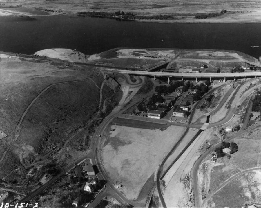 The new Interstate Highway 80 bridge that will span the entrance to the artificial bay that will be created by the U.S. Army Corps of Engineers' John Day Dam Reservoir on the Columbia River at Arlington, Oregon, will be placed in use in June 1965. Shown in the lower part of the picture is the 15-acre newly prepared business section created by the artificial fill to elevation above the reservoir. The buildings in the center of the picture are not scheduled for demolishing until May 1967; however, access to the present temporary business district will be practically cut off once traffic is diverted across the new Interstate 80 bridge. With creating the John Day reservoir, the water will inundate the present temporary business section occupied by the buildings, and the new Arlington will be provided with a quay-type waterfront. The huge fill riverward is being used as a disposal center for rock and earth being removed in the grading of Interstate Highway 80 along the south-shore cliffs.