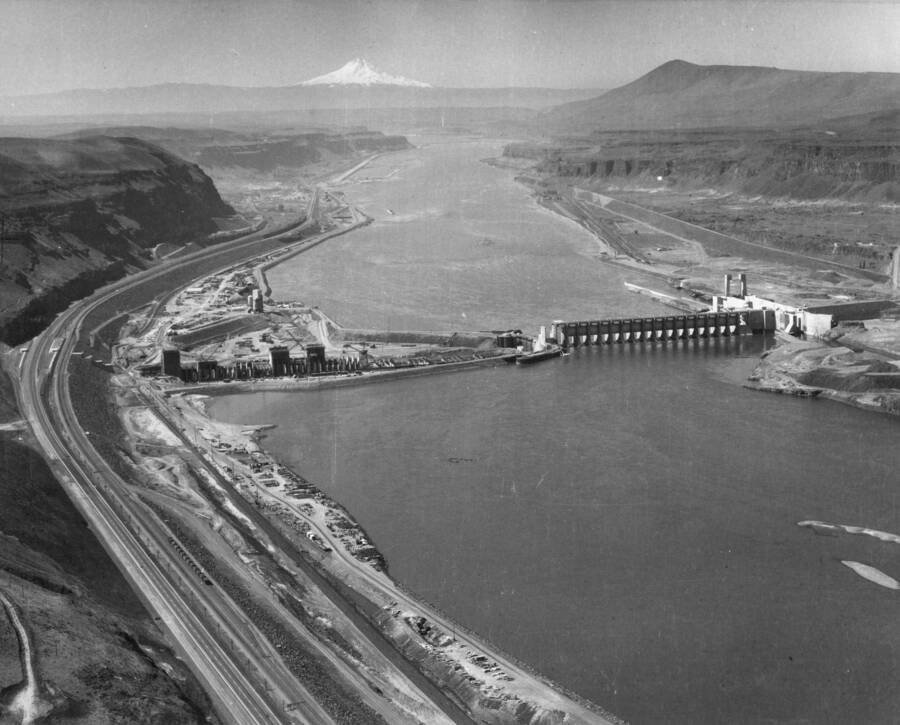 Looking downstream on the Columbia River, with Mount Hood in the background, is the U.S. Army Corps of Engineers John Day Lock and Dam. Work on the South or left shore, being carried on behind the 25-acre cofferdam enclosed work area, is nearing the 45 percent completion mark. Here the powerhouse and powerhouse intake section is being constructed, with a total of 578,000 cubic yards of concrete having been placed to date. Meanwhile, the entire flow of the Columbia River is being diverted through the 18 spillway bays while barge navigation is maintained through the North Shore navigation lock by use of a temporary channel feeding into the lock from upstream.