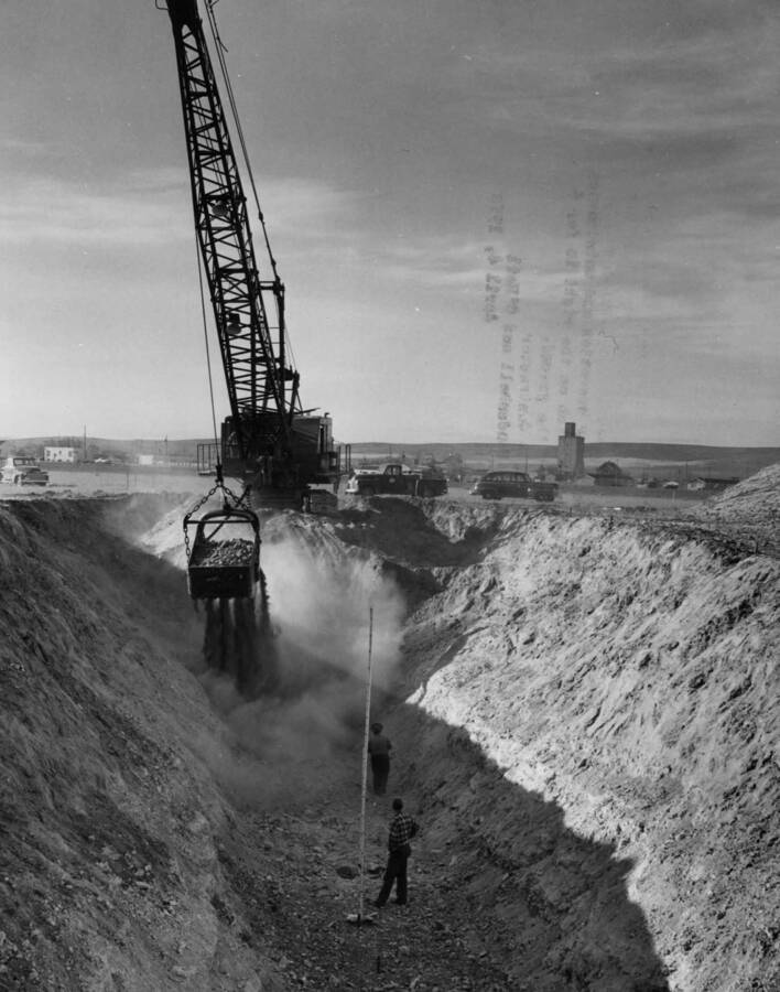 Columbia Basin Project, Irrigation Division, Third Section West Canal, Specifications 2844, Schedule 2. View looking north westerly into ditch being excavated for W 645 drain culvert under third section of the West Canal at station 1632.25. The buildings in the background are a portion of homes and businesses in the incorporated city limits of Quincy, Washington. Marshall, Haas and Royce and Haas and Rothschild--Contractor. H.E.Foss, Photographer.