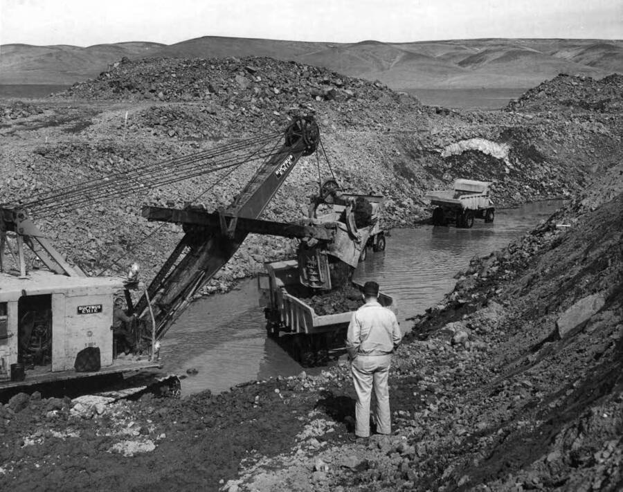 Columbia Basin Project, Irrigation Division, 2nd section West Canal, Specs. 2541. 54-B shovel is loading 10 yard Euclid trucks with rock from the G.N.R.R. crossing plug at the station 1043/20. Water in the canal came from snow melt and subsurface infiltration. A small patch of snow appears above the truck at the right. The subsurface flow had to be pumped out of the canal. Work is being performed by Morrison-Knudsen, Inc. under Schedule 2 of above specifications. H.E. Foss, photographer.