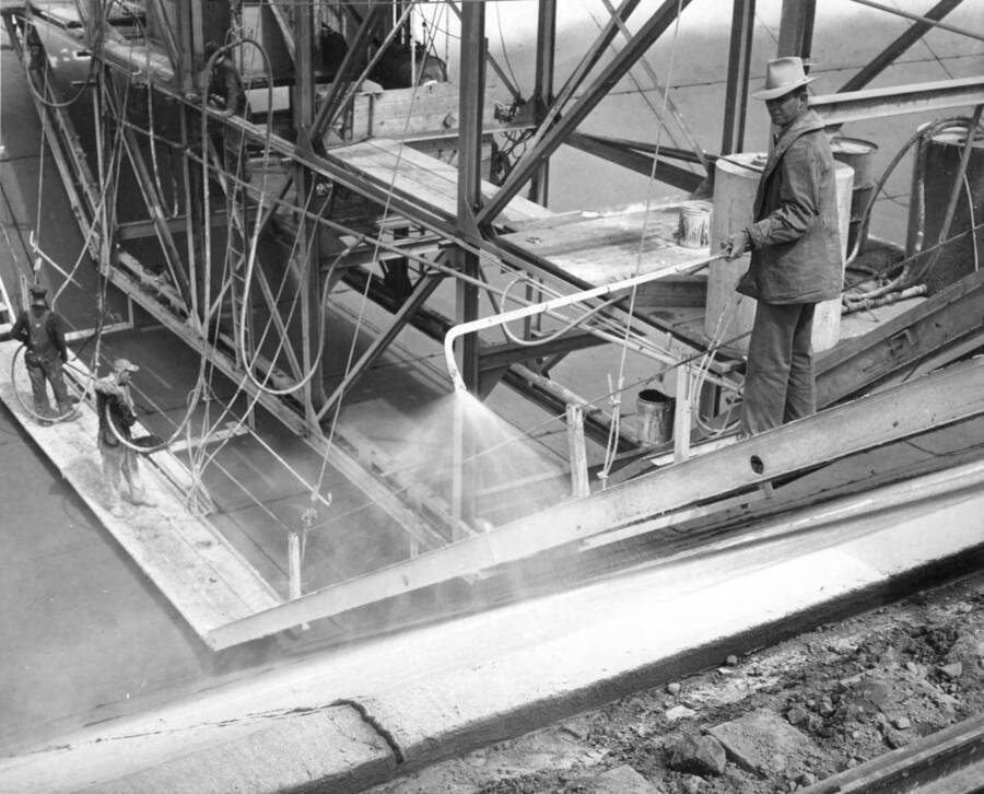 Columbia Basin Project, Irrigation Division, 2nd Section West Canal, Specs. 2541. Workman in the foreground is applying white pigmented curing compound. The two men on the platform in the bottom of the ditch are filling grooves with mastic. The man on the platform near the center of the machine controls the travel of the curing jumbo. Work is being performed by Morrison-Knudsen, Inc. under Schedule 2 of above Specifications. H.E. Foss, photographer.