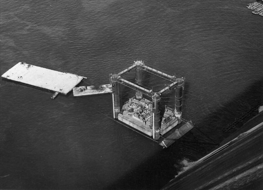 Specifications No. 2475, March 24, 1949. This photograph was taken as the floating caisson was being submerged in the spillway bucket during its initial trial operation. The caisson barge with operating equipment is shown floating between the caisson towers. In the foreground is the caisson 'A' frame anchored to the spillway face, and in the background are the divers' barge, the work barge, and the personnel skiff. This work is being performed by Pacific Bridge Company, as part of their contract for repair of the spillway of Grand Coulee Dam.