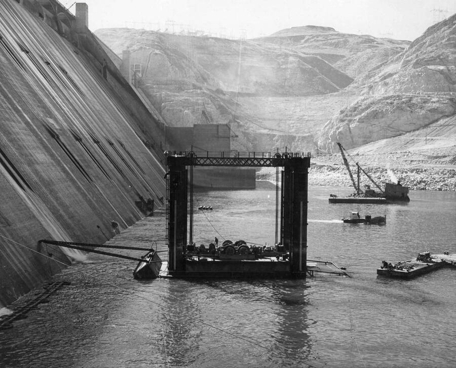 Specification No. 2475, March 24, 1949. When this photograph was taken the floating caisson was anchored by its 'A' frame to the spillway bucket section. The caisson barge with machinery for operating the caisson is between the towers. At the left are rafts carrying power line to the caisson barge. In the background are floats carrying maneuvering lines, a sea mule, and a dredging barge. This operation is being performed by Pacific Bridge Company, contractors, as part of their contract to repair the spillway bucket section of Grand Coulee Dam.