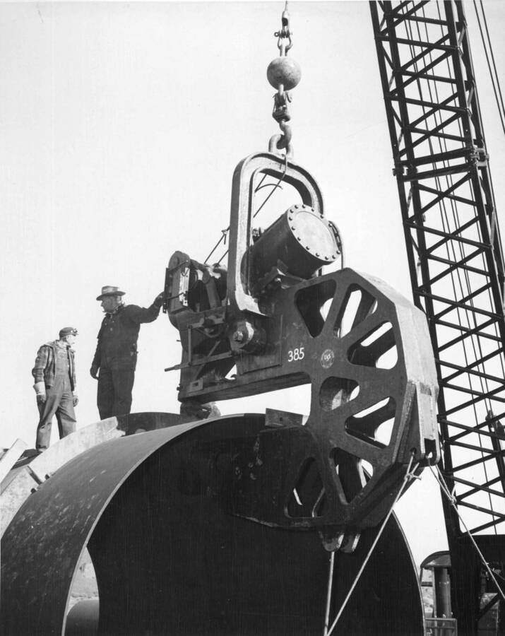 Specifications No. 2329. Picture Series 18532-3 through 18537-3. Morrison-Knudsen Co., Inc., and Peter Kiewit Sons' Co., subcontractor, Consolidated Western Steel Corp., Electric City Yard. A view of the process of mounting the stiffener rings on the discharge pipe with the aid of a compressed air-activated bull press. Rings are fabricated from quarter segments, with one joint being welded to the pipe as temporary lugs to keep the ring in alignment. 'The Bull' presses the ring into firm contact with the pipe and it is tack welded with a 4 inch fillet weld. The pipe is revolved approximately one foot by the positioning rollers, and the process is repeated until the ring is entirely tack welded. The pipe will then be rolled down the line to an automatic welder set-up (picture 18532-3) where a full fillet weld will be placed on both sides of the stiffener rings to complete the pipe for placing in the tunnel.