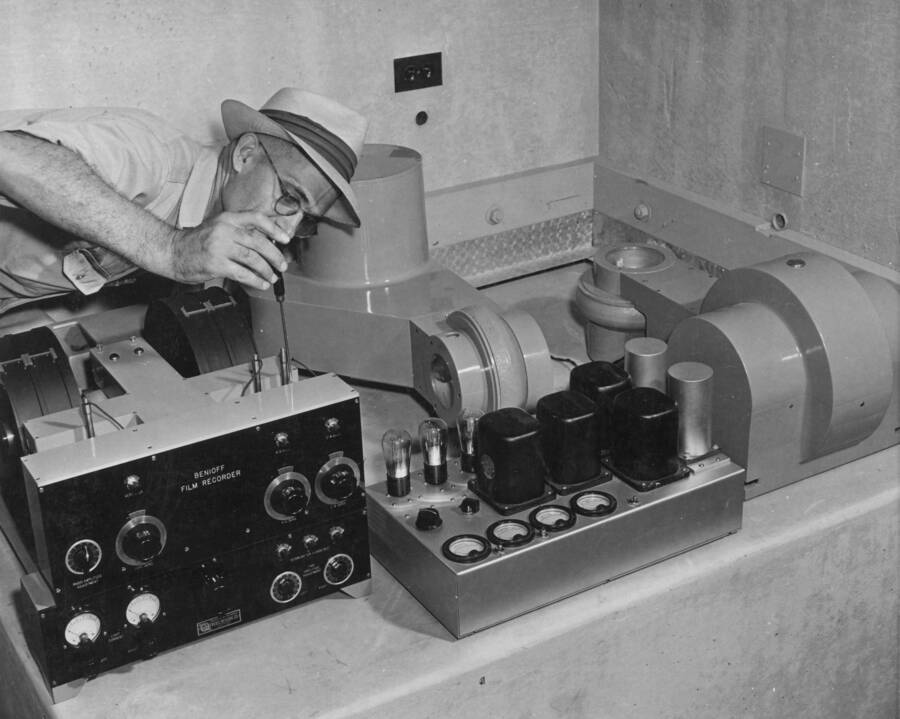 Grand Coulee Dam Seismograph Station, showing seismograph equipment before installation, arranged only for photographic purposes. Dr. Dead S. Carder of the U.S. Coast and Geodetic Survey is simulating the adjustment of position of galvanometers on the Benioff film recorder. In the right foreground is the 10-cycle tuning fork amplifier, and in the back, a horizontal and vertical seismometer