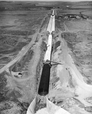 Columbia Basin Project, Irrigation Division, West Canal, Specifications 1286, Utah Construction Co. and Winston Bros. Co., Contractor. Looking east along the west canal showing Dry Coulee Siphon No. 2 in the foreground, the section of 12-foot bottom lined canal, Dry Coulee Siphon No. 1 and another portion of the lined canal in the background. A portion of the warped open outlet transition is shown in the immediate foreground is a 40-degree curve. Also shown in the foreground is the reinforcing steel for the uncompleted portion of the siphon barrel and the siphon form in place over a completed standard 25-foot section of the siphon barrel. The forms for the closed inlet transition are under construction. The contractor's shops and offices are on the right of the canal section. H.E. Foss, photographer.