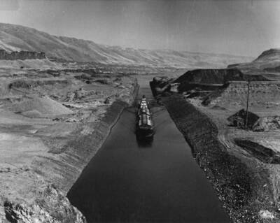 With diversion of the Columbia River through the North Shore spillway bays at the U.S Army Corps of Engineers' John Day Lock and Dam, barges and tows are now utilizing the temporary channel and permanent lock of the huge multipurpose project. Here is shown a tug with a pair of wheat barges in tow moving through the temporary channel, headed downstream for clearance through the John Day Lock. With completion of the project, this temporary channel will be inundated by the John Day Reservoir which will be nearly 100 feet in depth at this point.