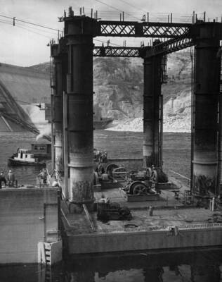 Floating Caisson aft section passing thru the drydock gates as it moves out into the drydock channel, where it will be moored while the Caisson Seat Frame is being assembled in the drydock. Foreground, Caisson with operating machinery on deck. Left, drydock well end miter gate. Background, tug and steel derrick barge. This work is part of the Pacific Bridge Co. contract, Specs. 2475, Coulee Dam, Washington.