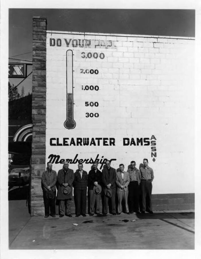 Some of the directors of the Clearwater Dams Assn. Standing in front of marker on theatre building, Orofino, showing goal of $3,000 which was not reached. Note on back: "" Some of the directors of Clearwater Dams Association Left to right: Frank Gaffney, L.G. Beeson,  , Mel Snook, Bob Oud, Dude Warren Gardner (city attorney),  Choate, Chuck Musial.""