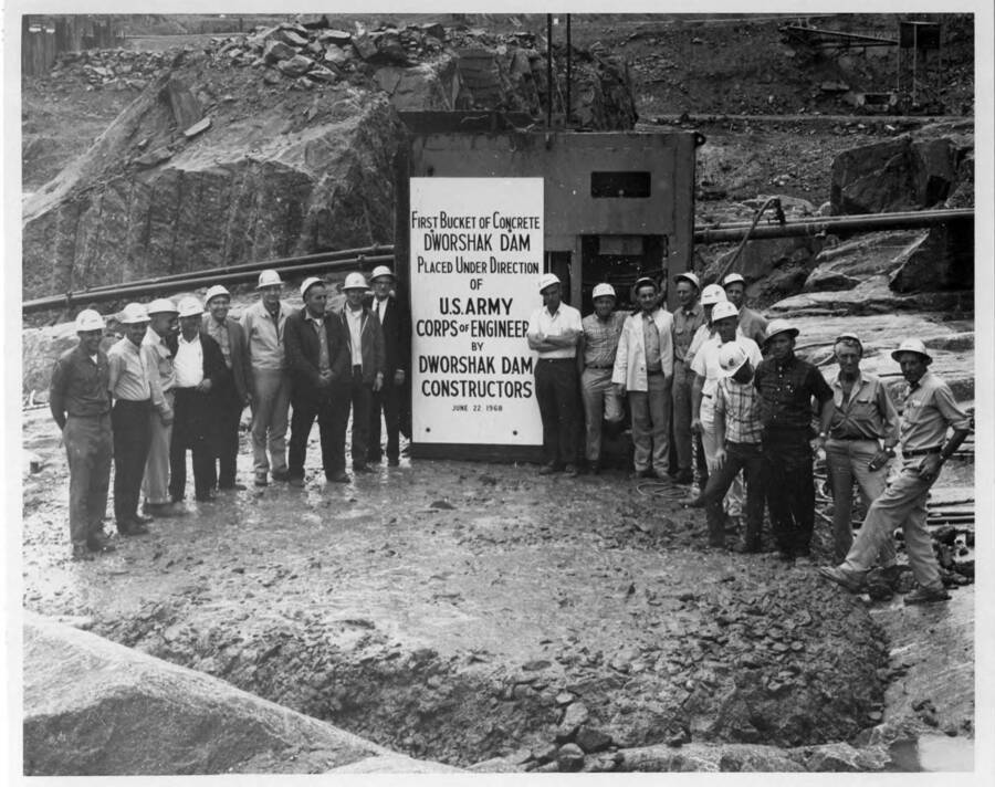 A gathering of the crew at the time of the first bucket of concrete poured at the project on June 22. Note on back: Left to right Carroll Johnson, Cableway Supt.; Dave Pruitt, Drill & Rock Supt.; Floyd Huntington, Cableway Supt.; Harold Taylor, Quarry Supt.; Hadley Garrison, Safety Supt.; Ed Hershberger, Project Engineer; John Bellegante, Shift Supt.; Ron Maxwell, General Supt.; Whitey Wixson, Project Manager; Bill Steffenson, Shift Supt.; Gene Jones, Carpenter Supt.; Pete Enevold, Electrical Supt.; Bill Little, Batch Plant Supt.; Bill McGee, Rigging Supt.; Vern Anderson, Master Mech.; Art Gress, Assist. Purchasing Agent; Jim Robertson, Plant Engineer; P.J. Parton, Pipe Supt.; Wayne Baldwin, Excavation Supt.; Ernie Meyers, Labor Supt.
