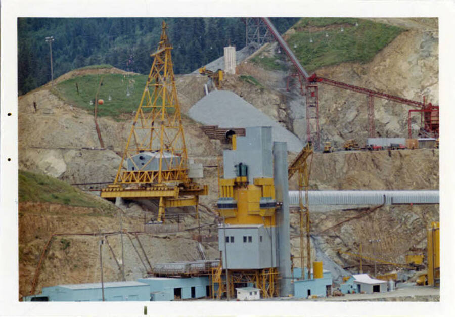 Photograph taken in May.  Shows top of the batch plant where they mixed the concrete.  Note on reverse reads 'Note one of the cable towers at the extreme downstream end of the track.  This would indicate that they were placing concrete on the very downstream edge of the dam.  Also seen is some of the crushed rock used for concrete -- the long tubes carries fly ash, a cement ingredient from Chicago that sets the concrete and makes it harder.'