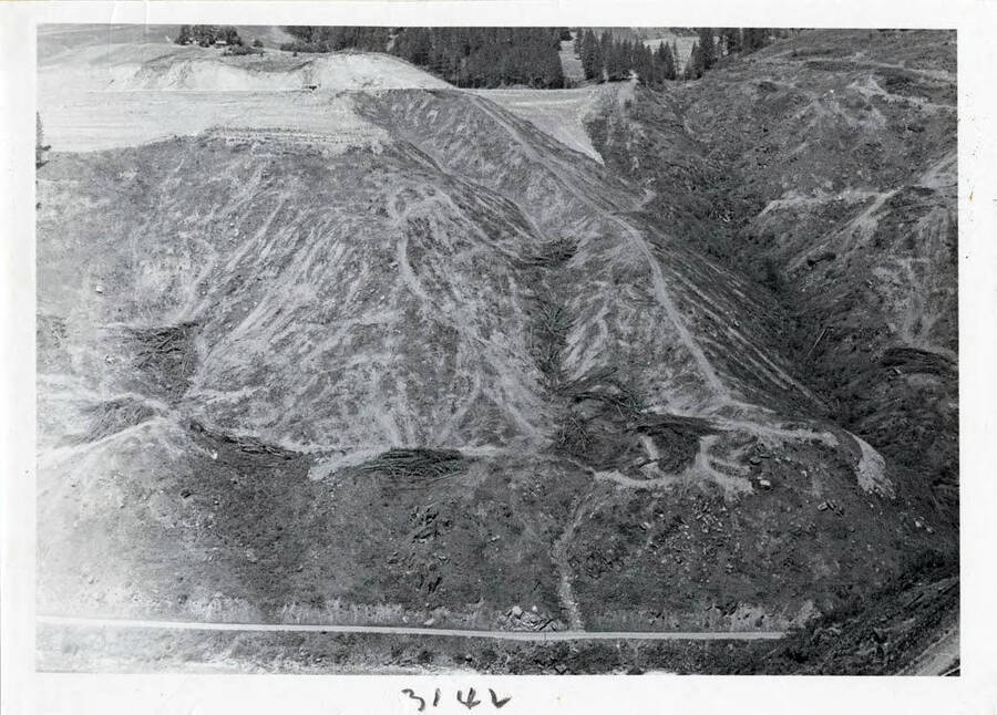 Photograph of clearing at the site to prepare for construction.  On top side you see fine new Army road completed into top of dam site and lower along the lower side of photo you see the original old road that went up the river.  It now goes to the base of the dam.  All material was taken from the site down to 2' in diameter and 2-3 ft. long.  Had to be burned or removed from area.
