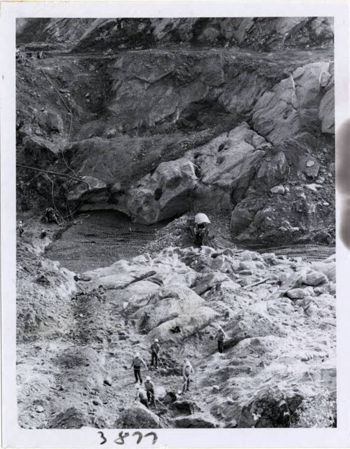 Photograph of the very bottom of the dam, elevation about 985 above sea level.  Here you see men cleaning up the bedrock and solid granite just as you would clean the kitchen floor.  It is at this very spot that the concrete started to pour into the base.  You can see the clean granite formation, base of dam, just before the concrete was put in.