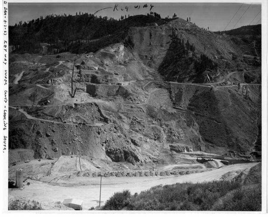 Work just starting at the dam location. Note on image: "" D Dam - 8-3-67 Key way under cons't - looking south."" Note on back of image: ""You will note where marked that they have blasted and skimmed off the dirt and loose rock. This is to be the 'Key Way' or axis of the dam. By putting these photos together with the original one of this set you can see just about the location. Looking south. Also note the coffer dam is being built and the diversion tunnel partly in. Taken August 3, 1967.""