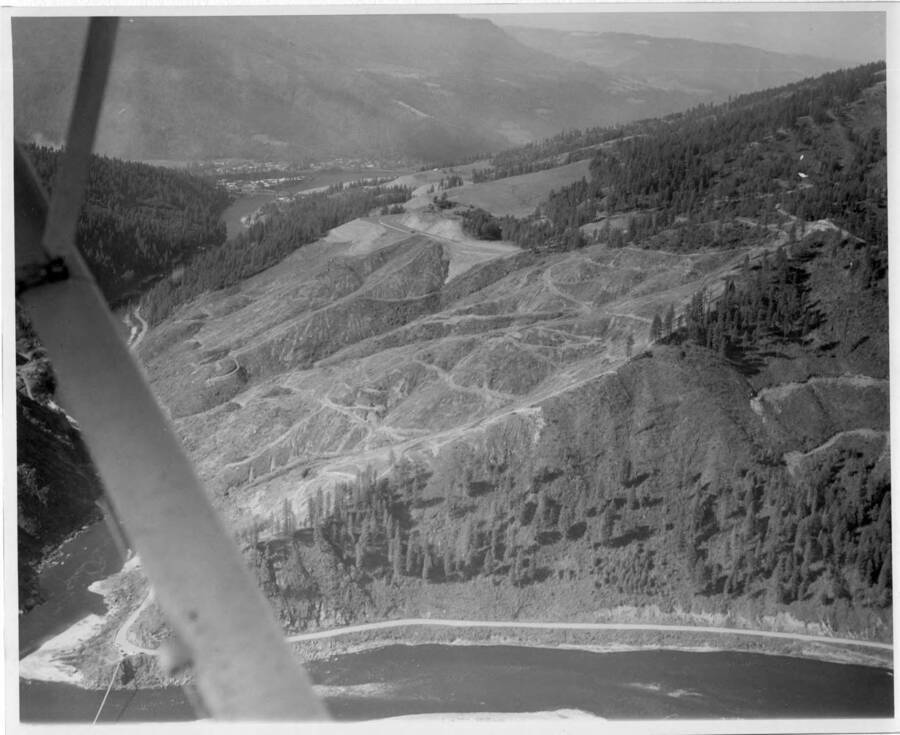 Strip work underway.  Note on back: ""Aerial view. Strip work underway and here you will see the new access road on the south side of the river built by Murphy Bros. of Spokane. This comes into the upper side of the dam, for construction. Riverside-Orofino far downstream, 1.5 miles.""