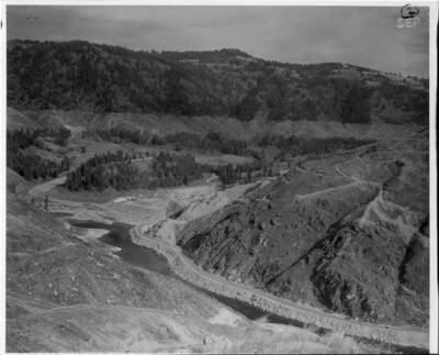 During early construction stages a road was built up the south side of the river to the contractors shops location. Note on back: "" Land clearing has started at elevation 1605 on the distant hills. This will be the pool level.""