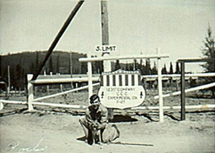 No legible name or date. Man stands with a dog in front of the entrance sign to the CCC camp located on Big Creek.