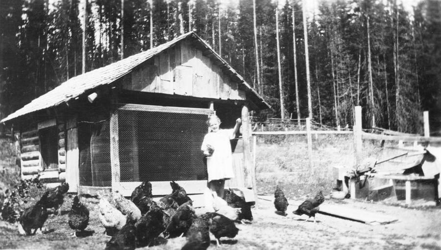 Caption titled: "Margaret Elaine Larsen feeding chickens, Priest River Experimental Station, Priest River, Idaho". Old site maps place a chicken coop in the vicinity of the present fuel house.