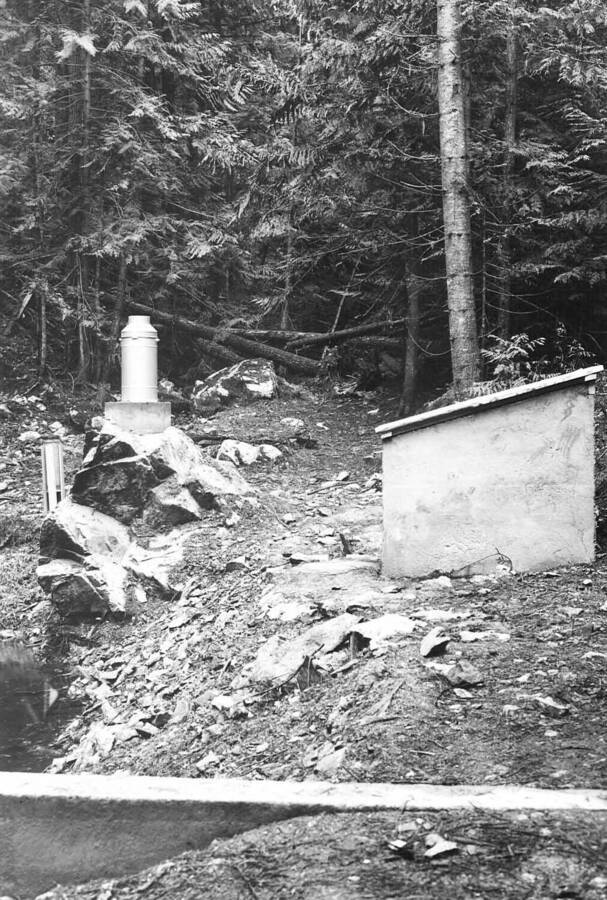 Back of photo reads: "Weather Bureau standard rain gage, Friez recording rain and snow gage (on rock), and Stevens water-level recorder gaging house. Benton Creek stream-flow gaging station, Priest River Experimental Forest. November 20, 1939. f-22, 1 sec., distance at 30 ft., verichrome film. DM 39-1."