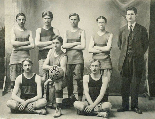 Eight young men are posing for a group portrait. Seven are wearing sleeveless athletic uniforms, with five standing and two seated cross-legged in front. One man, dressed in a suit, stands at the far right. The man seated in the center is holding a basketball, which has "1924 OSA Class A Champs" written on it.