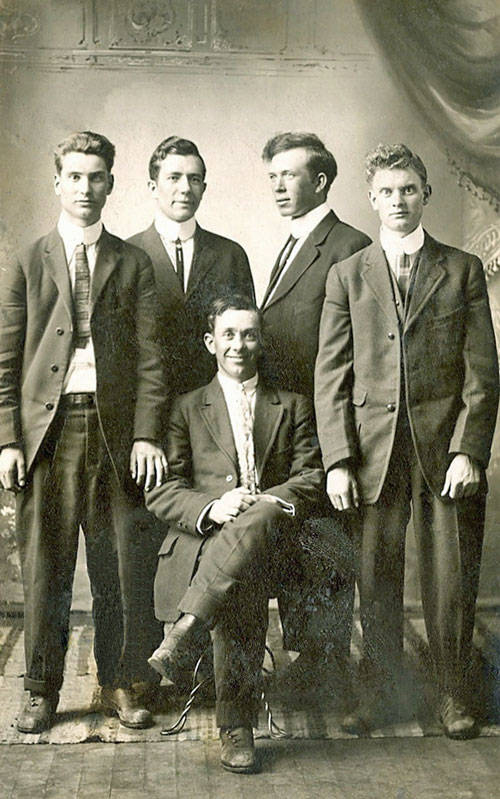Five men in suits pose for a formal group portrait. Four stand in the back row, and one sits in front of them with his arms crossed. The background shows a draped curtain and ornate wall detailing.