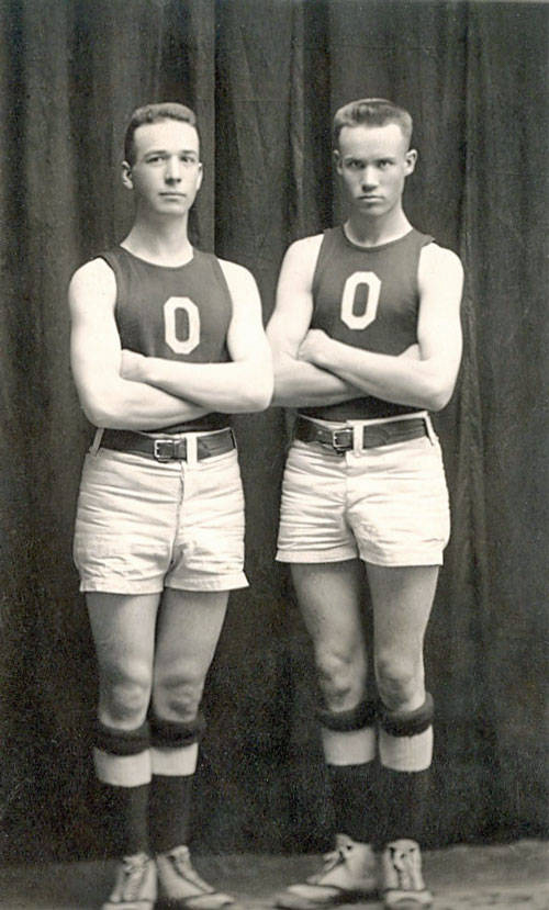Two young men are standing with their arms crossed, wearing athletic uniforms. Each uniform consists of a sleeveless top with the letter "O" on the front, paired with belted shorts, high socks, and athletic shoes. The background is a plain, dark curtain.