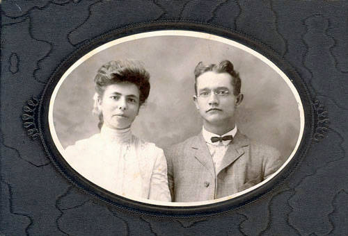 A woman and a man are posed side by side in an oval frame. The woman has her hair styled up and is wearing a high-collared dress. The man is wearing glasses, a suit, and a bow tie. The background is plain, and the portrait is set against an embossed decorative border.