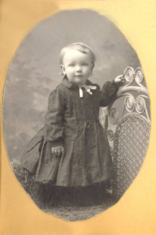 A young child in a long dress standing beside an ornate wicker chair, with a neutral backdrop behind.