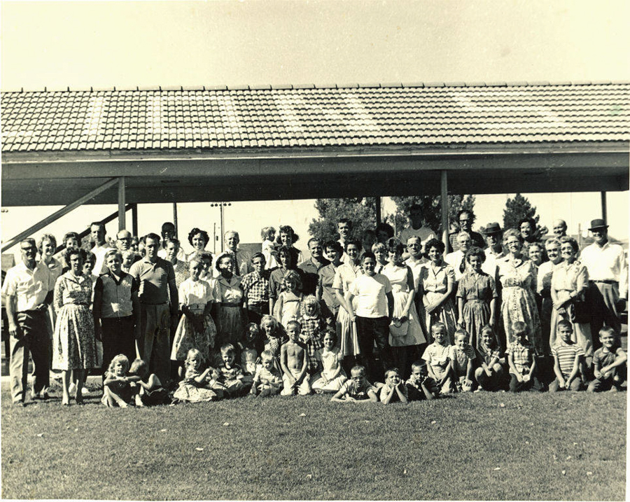 A large group of adults and children pose together on grass in front of a building with a tiled roof. The adults stand in the back and the children sit in the front. The scene is outdoors with trees and a clear sky visible in the background.