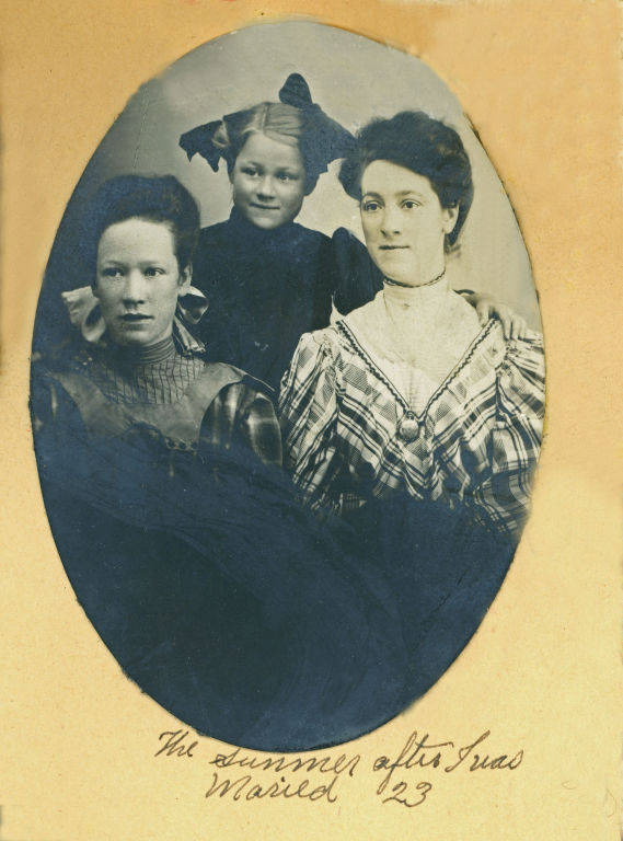 Three women posed for a portrait. Two women are seated, and a younger girl stands behind them. The woman on the left wears a dark dress, while the one on the right is in a patterned dress with high collar. The young girl has bows in her hair. Handwritten text below reads, "The summer after I was Married ’23."