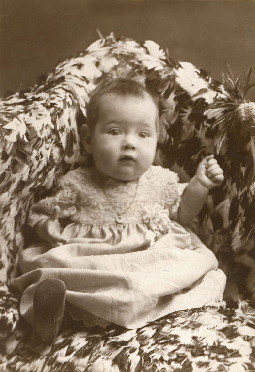 A baby wearing a lace dress is seated on a patterned blanket, looking forward with one hand raised.