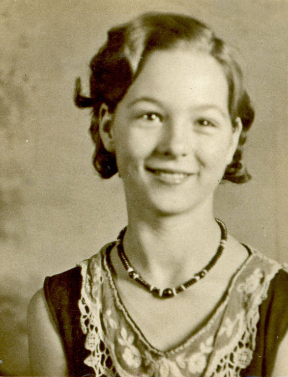 A young woman with short, wavy hair smiles at the camera. She is wearing a dress with lace details and a beaded necklace. The background is plain.
