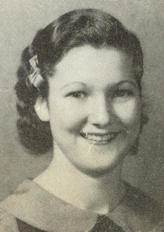 A woman with short wavy hair, wearing a blouse with a collar. She is smiling and has a small hair accessory on the side.